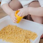 A child filling a yellow cup and playing with a sensory tub filled with dry elbow macaroni noodles.