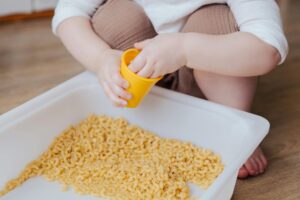 A child filling a yellow cup and playing with a sensory tub filled with dry elbow macaroni noodles.